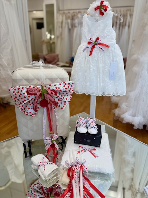 White lace baby dress on a stand with red ribbon bows, surrounded by coordinated accessories and white quilted luggage with a red bow.