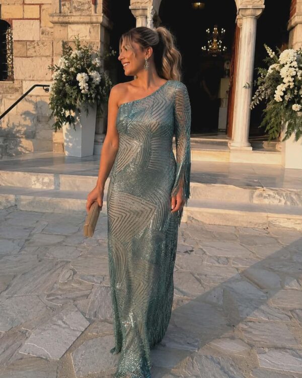 Woman in a shimmering teal, one-shoulder sequined gown with a fringed sleeve posing on stone steps outside a historic building with floral arrangements in the background.