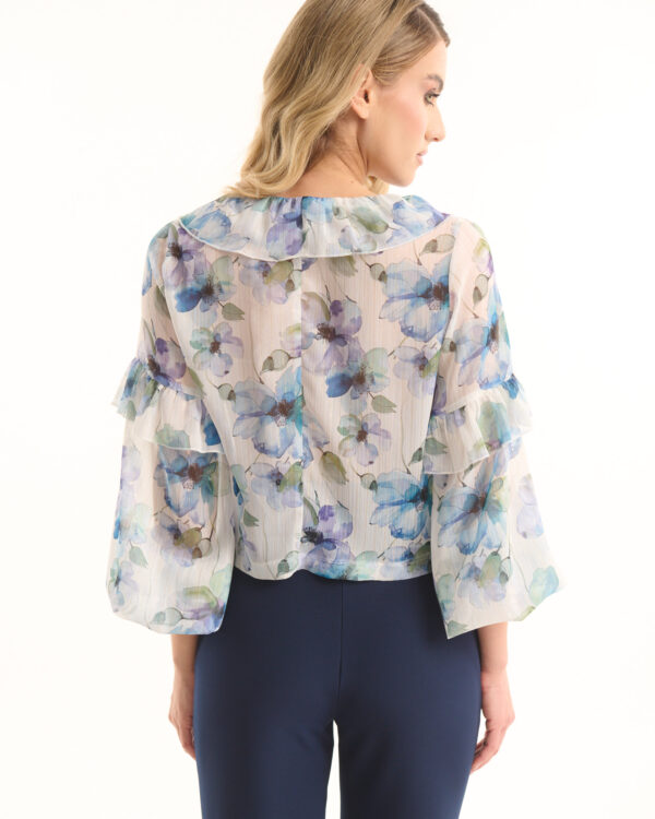 Back view of a woman wearing a sheer floral blouse with blue and purple flowers and ruffled sleeves, paired with navy pants.