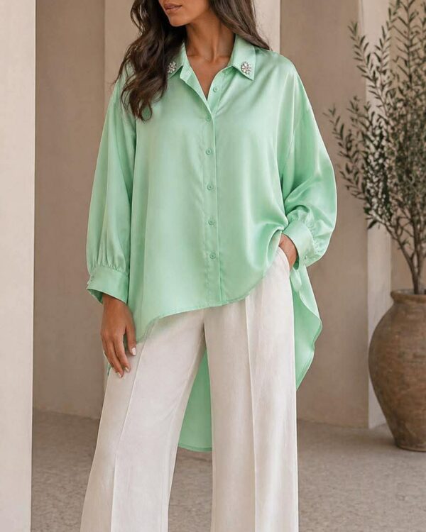 Model wearing a mint green oversized blouse with white wide-leg pants, standing indoors near arches in a minimalist setting.