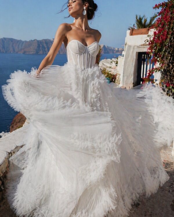 Bride in a white strapless corset gown with a voluminous layered tulle skirt standing on a seaside terrace, breeze blowing her dress.