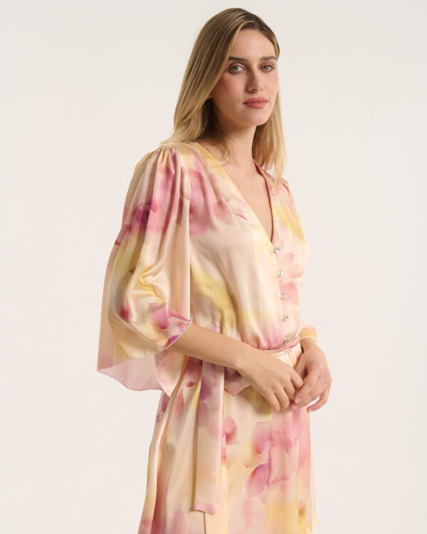 Woman wearing a flowing pastel floral dress, posing against a pale background and looking at the camera.