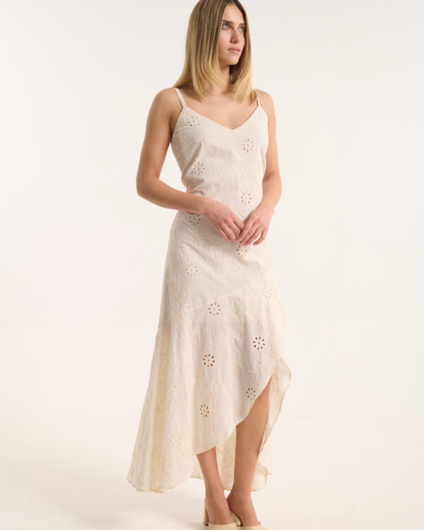 Full‑length image of a woman in a cream floral dress with a slit, standing against a light background, wearing beige sandals and looking to the side.