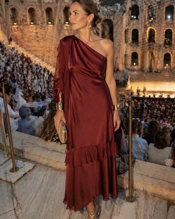 Woman in a burgundy one-shoulder satin dress with ruffles, gold jewelry, and a small metallic clutch posing in an ancient amphitheater filled with spectators at dusk.