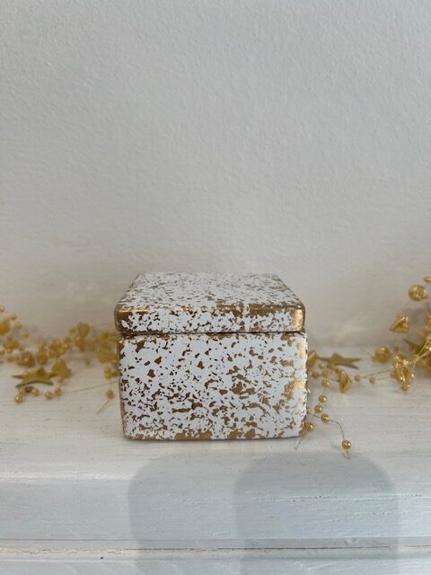 Small white gift box with a gold speckled, distressed finish sits on a white mantel with gold beads in the background.
