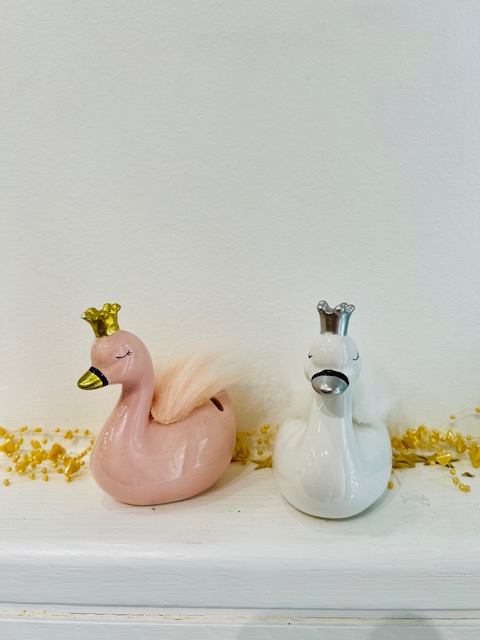 Two ceramic swan figurines, pink and white, wear metallic crowns and sit on a white surface with gold beads nearby (decorative display).