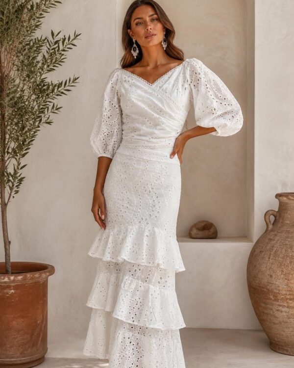 Woman in a white eyelet lace midi dress with puff sleeves and a tiered ruffle skirt, posing in a sunlit courtyard.