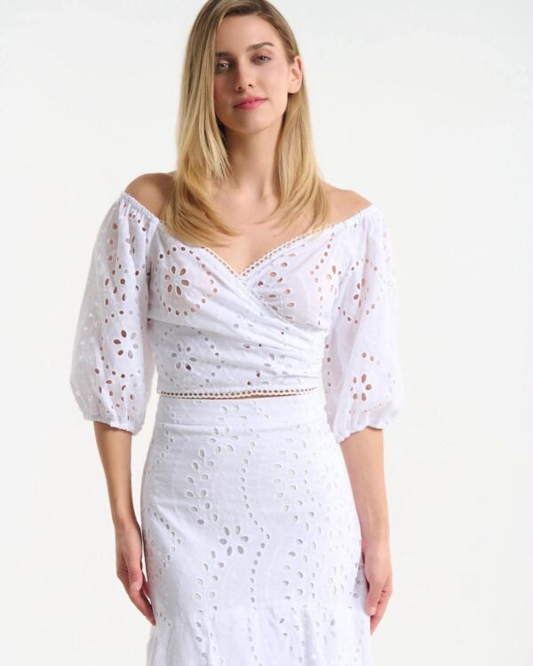 Woman wearing a white off-the-shoulder eyelet dress with puff sleeves and a knee-length skirt, standing against a light background.