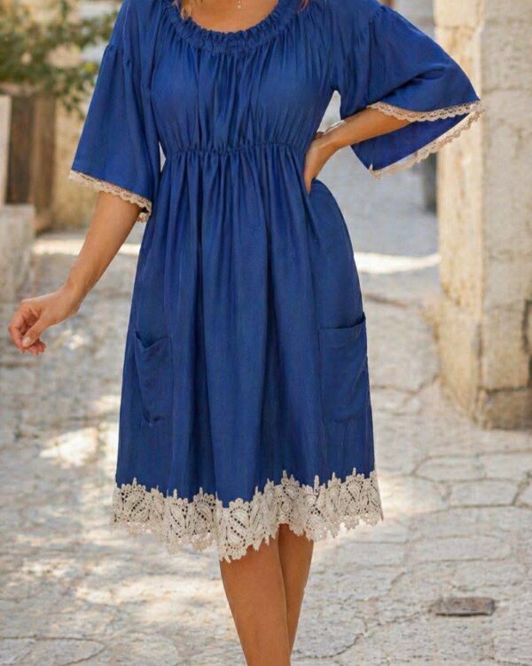 Model wearing a blue knee-length dress with white lace trim on sleeves and hem, standing on a sunlit stone street with pockets visible and sandals.