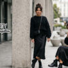 Urban fashion scene: two women in black outfits on a city sidewalk, one standing by a concrete pillar with a crossbody bag, the other seated with a black handbag; Miss Vanity logo on the left.