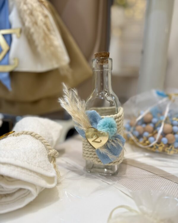 Small glass bottle with a cork stopper, wrapped in rope with blue ribbon and a gold heart charm; decorative items on a table in the background.