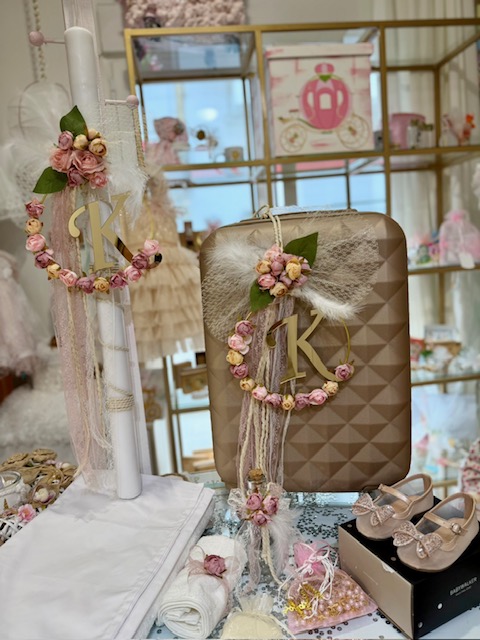 Decorative boutique display featuring a pink floral wreath with a gold ‘K’ on a white pillar and a matching geometric beige case, with pastel accessories around it.
