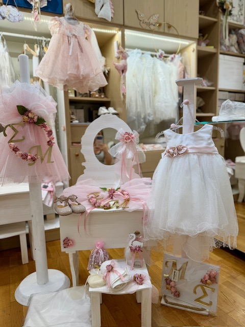 Pastel pink and white children's dresses and accessories arranged on a display in a princess-themed boutique, with a round mirror and floral accents.