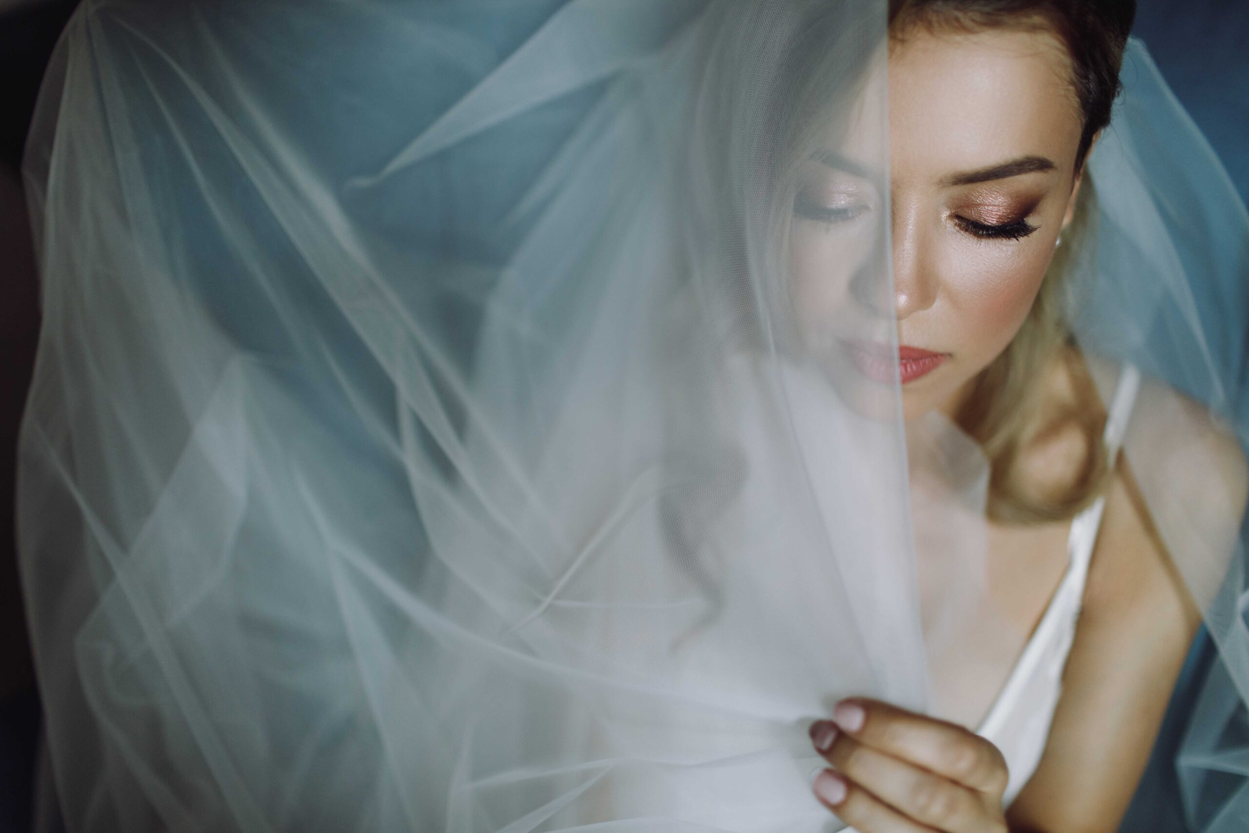 Woman with a sheer veil draped over her face, eyes closed, makeup visible, soft lighting (bridal portrait)