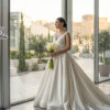 Bride in a long white wedding gown holding a bouquet of white calla lilies in a glass-walled atrium; Miss Vinity logo visible in the corner.