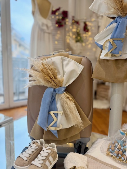 Brown suitcase decorated with white and blue ribbons, dried pampas grass, and a gold lambda charm in a cozy, well-lit room setting.