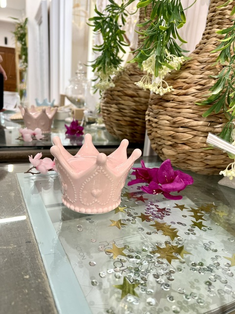 Pink ceramic crown ornament on a glass display table, surrounded by greenery and scattered gold star confetti.