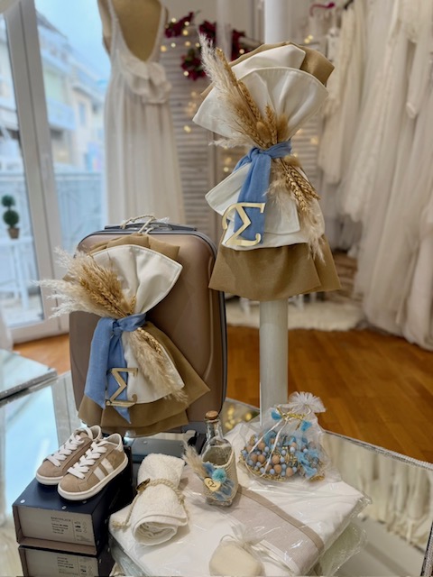 Wedding shop display of coordinated bridal accessories: a tan suitcase and a dress form decorated with beige and blue ribbons and dried pampas grass, with small shoes and favor bags on a glass table in the foreground.