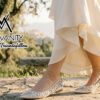 Miss Vanity logo over a model wearing bejeweled beige flats walking on a sunlit stone path in a flowing cream dress with greenery in the background.