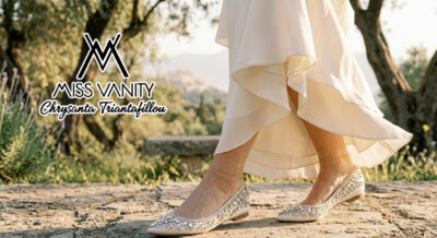 Miss Vanity logo over a model wearing bejeweled beige flats walking on a sunlit stone path in a flowing cream dress with greenery in the background.