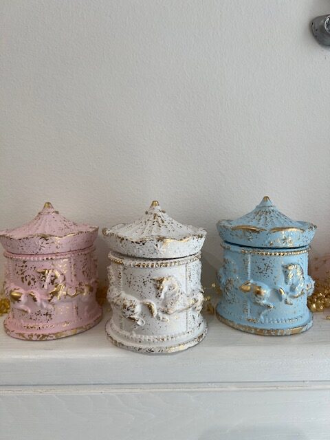 Three pastel, ornate ceramic jars with domed lids and gold distressing lined up on a white shelf (pink, white, blue).