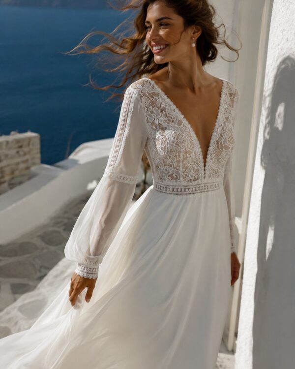 Bride in a white lace wedding dress standing on a sunlit seaside terrace, smiling at the camera with the ocean in the background.