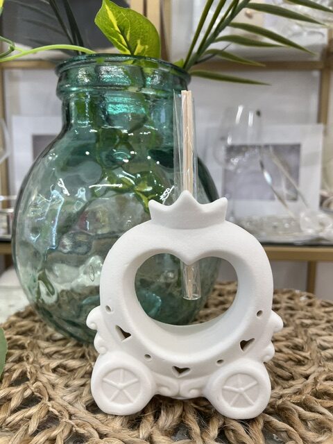 White ceramic carriage-shaped ornament with a crown top and circular center, placed in front of a teal glass vase on a woven surface.