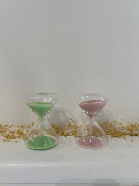 Two glass hourglasses with green and pink sand sit on a white surface, with gold bead garland in the background (decorative timer scene).