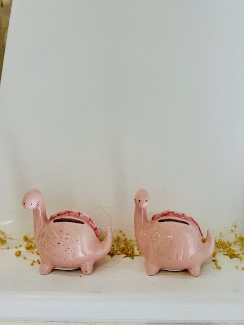 Two pink dinosaur-shaped piggy banks with coin slots on their backs sit on a white shelf with gold bead confetti nearby.
