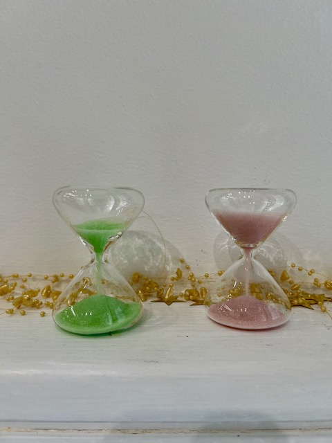 Two glass hourglasses on a white surface, green sand in the left one and pink sand in the right, with gold confetti in the background.