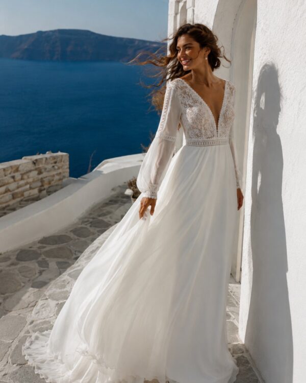 Bride in a white lace-eluce wedding dress standing by a white wall with a flowing skirt and lace bodice, sea in the background