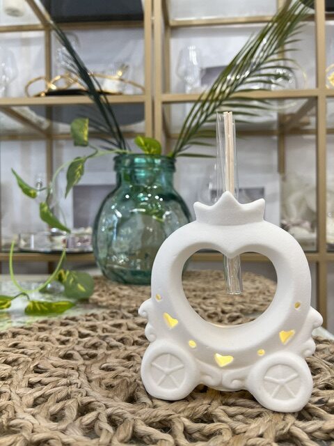 White donut-shaped decorative candle holder with a straw in the center, on a woven mat, beside a green glass vase and plants in the background.