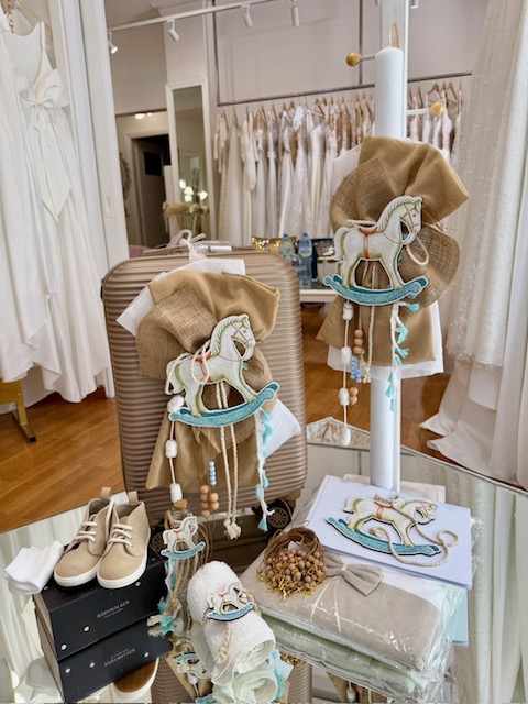 Rustic wedding decor on a glass table: burlap hoops with embroidered rocking horses, beads, and teal accents, beside a beige suitcase and shoes in a bridal shop.