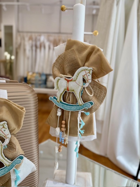 White taper candle on a stand wrapped with burlap and a fabric carousel-horse ornament, with tassels and beads hanging nearby.