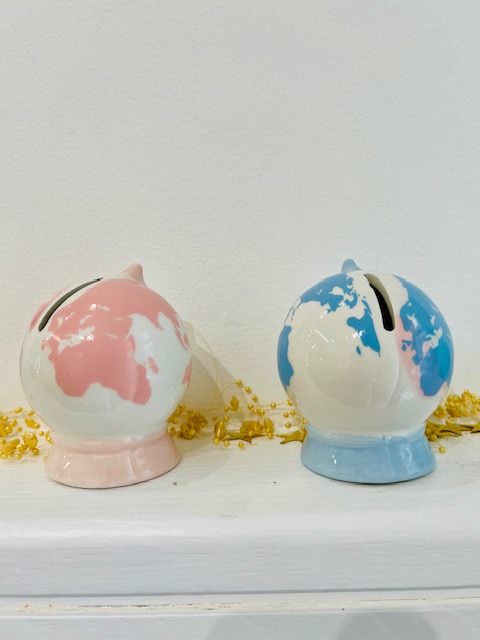 Two globe-themed piggy banks, pink and blue, sit on a white shelf with gold coins scattered around.