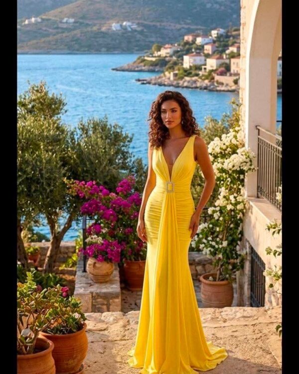 Woman in a yellow belted evening gown standing on a stone terrace by the sea, with flowering pots and a coastal town in the background.