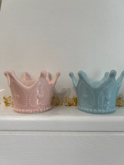Two pastel crown-shaped ceramic figurines, pink and blue, on a white shelf with a gold garland nearby