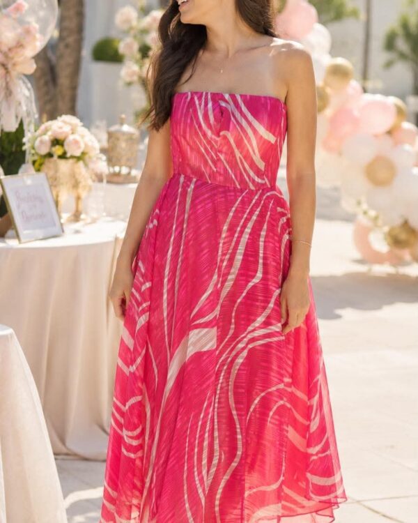 Woman in a pink strapless midi dress smiles at an outdoor celebration with pastel balloons and a decorated table in the background.