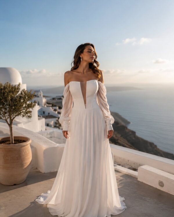 Woman in a white off-the-shoulder gown with sheer long sleeves stands on a terrace overlooking the sea at sunset.