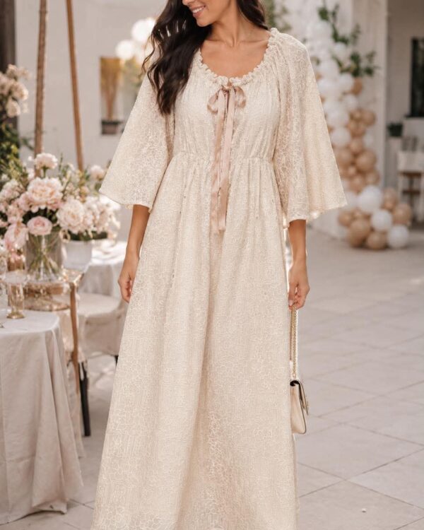 Woman wearing a cream lace maxi dress with a pink satin tie, standing at an outdoor decorator setup with flowers and balloons in the background.