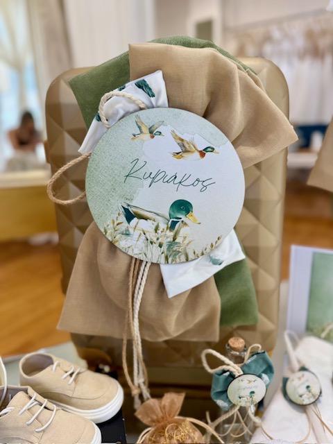Gift display with beige and green fabric draped over a textured box; round tag shows birds and the word 'Kupakos' attached by twine, with beige shoes visible at left.