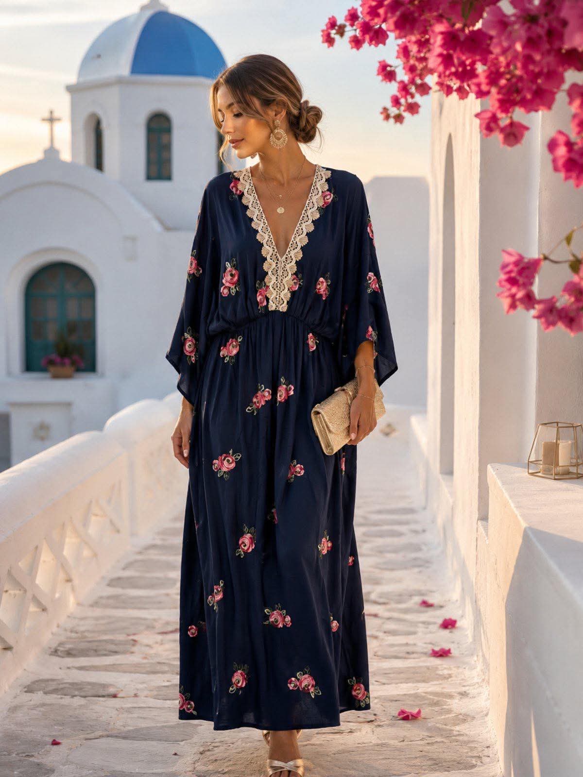 boho maxi forema me kenthtes leptopereies kai dantela(1) Woman in a navy floral maxi dress with lace neckline walks along a white stone path, holding a beige clutch, with pink flowers nearby and a blue-domed church in the background