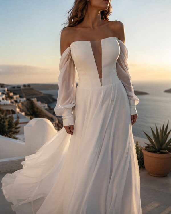 Bride in a flowing off-shoulder white wedding gown with sheer sleeves, standing at sunset overlooking a coastal town and sea.