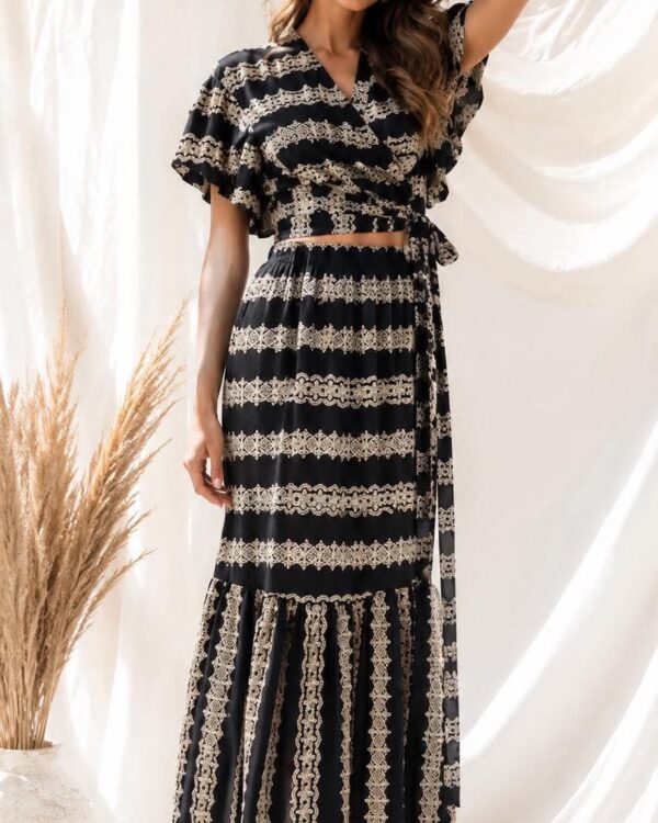 Woman wearing a black and beige lace two‑piece maxi dress, posing with one hand in her hair against a light draped backdrop, missvanity.gr text above (brand banner).
