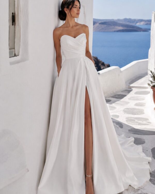 Bride in astrapless white gown with a high slit, leaning against a white wall by the sea with cliffs in the distance