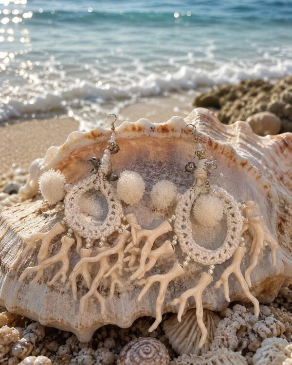 Cream crocheted hoop earrings with white pom-poms and coral-like beads, placed on a large seashell at the beach.