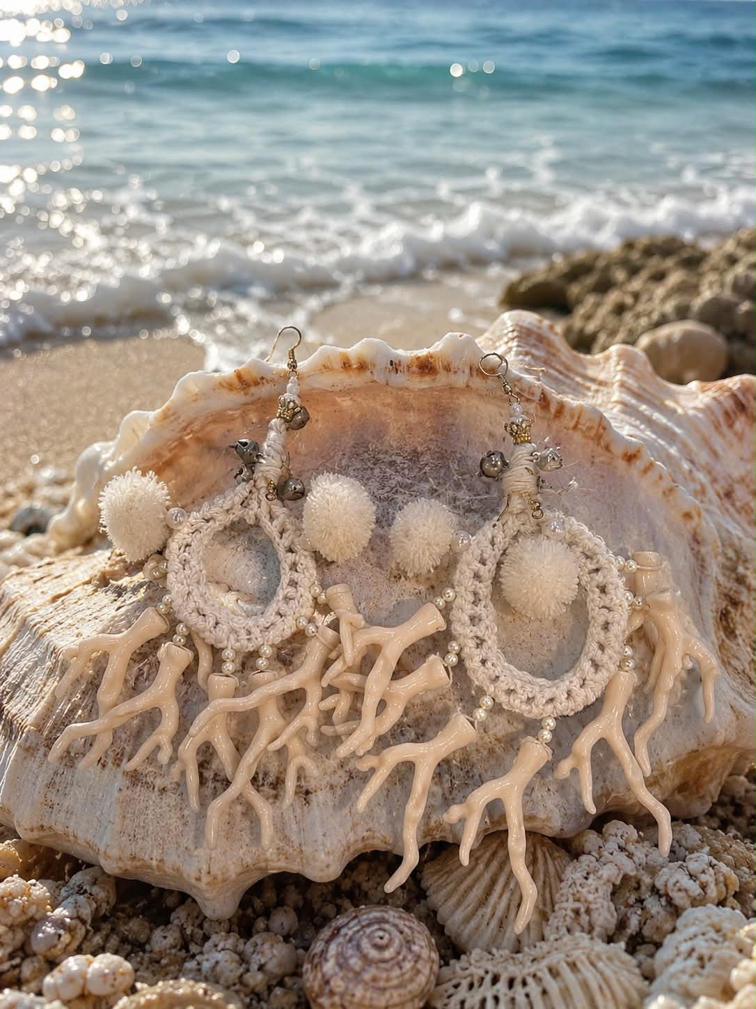 xeiropoihta plekta skoylarikia me korallia(1) Cream crocheted hoop earrings with white pom-poms and coral-like beads, placed on a large seashell at the beach.