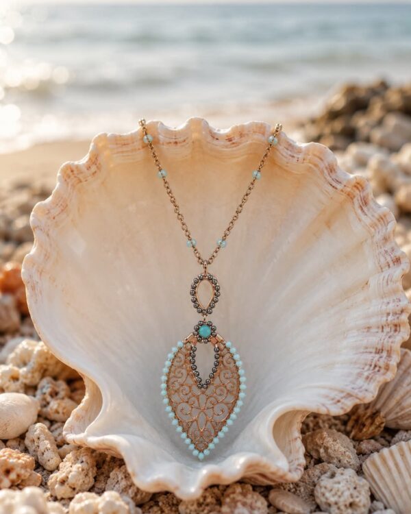 Gold-toned pendant necklace with turquoise beads resting inside a large seashell on a sandy beach, ocean in the background.