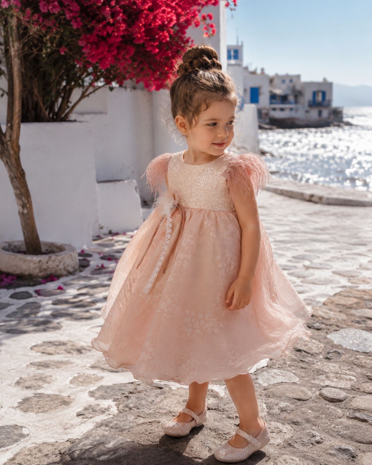 oloklhrwmeno paketo baptishs me thema to arkoudaki(3) Girl in a pale pink, sparkly dress with feather sleeves stands on a sunlit cobblestone street by the sea, looking to the side.