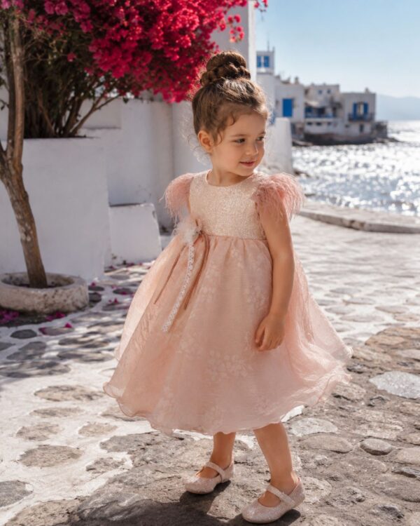 Young girl in a pale pink, sparkly dress stands on a sunlit stone street by the sea, with white buildings and pink bougainvillea in the background.